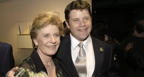 A medium shot of Sean Astin and Patty Duke smiling warmly while posing together at an indoor event. Patty Duke, on the left, has short blonde hair and wears a patterned blouse, while Sean Astin, on the right, is dressed in a dark pinstripe suit with a patterned tie