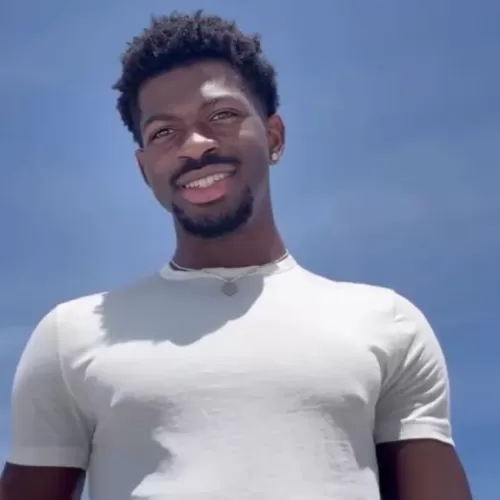 A low-angle shot of Lil Nas X smiling brightly at the camera in a white t-shirt against a clear blue sky, an image accompanying articles about a Lil Nas X bipolar diagnosis