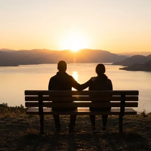 Silhouettes of two people sitting together on a wooden bench overlooking a calm lake and mountains at sunrise. One person rests a comforting hand on the other's shoulder, illustrating the power of connection and community support in breaking the stigma of bipolar disorder.
